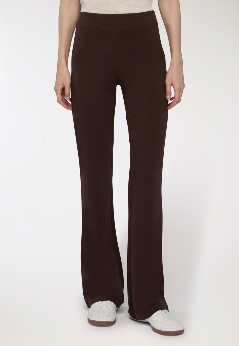 Osoba w ciemno-brązowych prostych spodniach i białych trampkach, stojąca na tle gładkiej białej ściany.