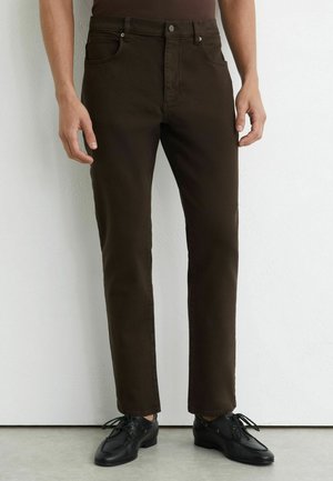 Person iført mørkebrune slim-fit jeans og sorte læder snørestøvler, stående op ad en hvid væg.