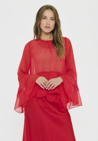 Rote durchsichtige Bluse mit Rüschensäumen über einem einfarbigen roten Rock. Leichter Stoff mit weicher Textur und rundem Ausschnitt.