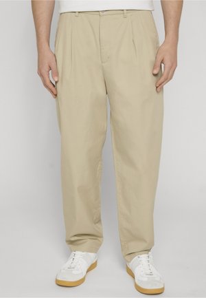 Mann trägt beige plissierte Hose und weiße Sneakers, steht vor einem einfarbigen hellgrauen Hintergrund.