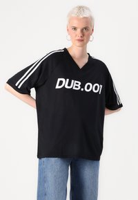 Melnā lielā t-kreklā ar V-veida izgriezumu, uz kura ir balti uzraksti "DUB.001" un balti svītriņas uz piedurknēm. Mīksta auduma, brīva piegriezuma.