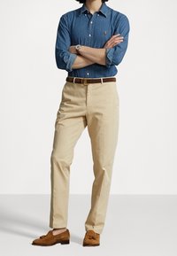 Camisa de mezclilla azul con botones, pantalones chinos caqui, cinturón de cuero marrón y mocasines de ante marrón con borlas, posando frente a un fondo neutro.