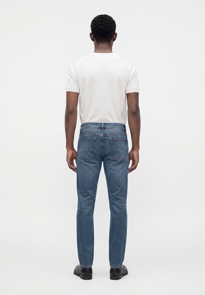 Homme debout de dos portant un t-shirt blanc à manches courtes, un jean bleu et des chaussures noires sur un fond clair uni.