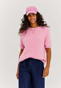 Ružový chlpatý sveter s krátkymi rukávmi, doplnený tmavými širokými džínsami. Modelka má na sebe zodpovedajúcu ružovú čiapku a vrstvené náhrdelníky. Viditeľná mäkká textura.