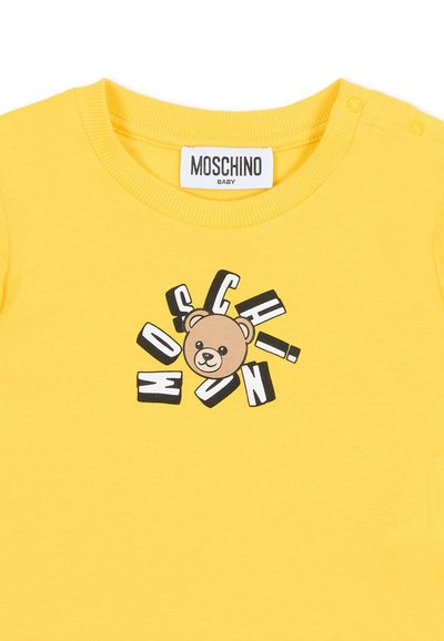 Camiseta de algodón amarilla con un gráfico de un oso de dibujos animados en el centro, rodeado por el texto "Moschino" en letras negras y blancas en negrita.