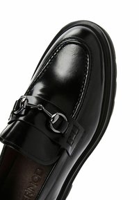 Mocasines de cuero negro con un acabado brillante, que presentan un detalle de hebilla decorativa, costuras elevadas a lo largo del borde y una suela texturizada.