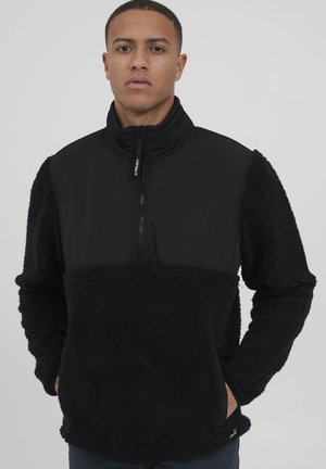 Giacca pullover nera con texture in fleece, chiusura a metà zip e colletto rialzato. Presenta un tessuto liscio a contrasto sulla parte superiore del petto.