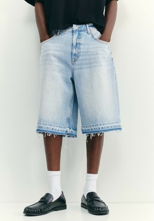 Personne portant un short long en jean bleu clair avec des ourlets effilochés, des chaussettes blanches, des mocassins noirs et un haut noir avec les mains dans les poches.