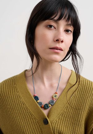 Cardigan en tricot moutarde avec un décolleté en V profond, orné d'un collier de perles multicolores avec des motifs floraux et un fil bleu. Fermeture simple par bouton.