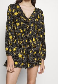 Vestido negro con estampado floral que presenta flores amarillas y lunares blancos, de mangas largas, escote en V profundo, y cintura elástica con detalle de lazo.