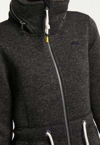 Mörkgrå zip-up jacka med hög krage, vita dragsnören med färgade ändar och liten paraplylogga på bröstet.