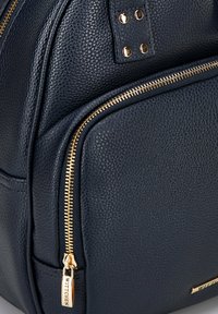 Sac à dos en cuir texturé bleu marine avec une fermeture éclair dorée en évidence, une poche latérale avec une tirette dorée, et des clous métalliques sur le rabat.