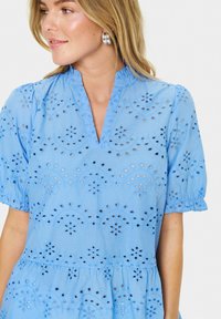 Blusa azzurra chiara con dettagli in pizzo ricamato, dotata di scollatura a V, maniche a palloncino corte e vita arricciata. Tessuto dalla texture liscia.
