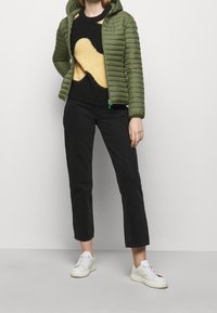 Doudoune verte avec capuche, présentant un design matelassé horizontal. Pull en tricot noir avec un motif abstrait jaune, pantalon noir fuselé et baskets blanches.