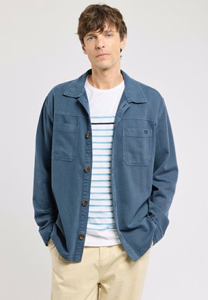 Homme aux cheveux bruns portant une veste bleue boutonnée sur une chemise blanche à rayures bleues et un pantalon beige, debout les mains dans les poches.