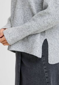 Hellgrauer Strickpullover mit einem lockeren Schnitt, seitlichen Schlitzen und gerippten Bündchen, kombiniert mit dunkelblauen Jeans. Weiche Textur, lässiges Design.