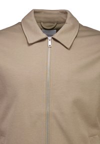 Beige vetoketjullinen takki, jossa on jämäkkä kaulus, metallinen vetoketju ja sileä kangaspinta. Minimalistinen malli ilman lisäkoristeita.