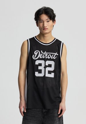 Giovane uomo con capelli neri e ondulati che indossa una canotta da basket nera a rete con la scritta bianca "Detroit 32", in piedi davanti a uno sfondo grigio semplice.
