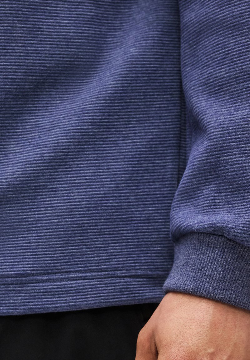 Gros plan sur la main d'une personne posée à côté d'un poignet de chemise bleue à manches longues texturée côtelée, au-dessus d'un pantalon noir.