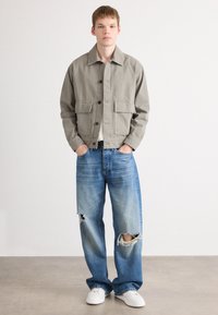 Lys grå lerretsjakke med to frontlommer, knappelukking. Brukt med slitte lyseblå jeans og hvite sneakers.