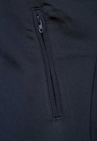 Laivastonsininen vetoketjullinen tasku joustavasta kankaasta valmistetussa vaatteessa, jossa on sileä pinta ja litteät saumat virtaviivaisen ulkonäön takaamiseksi.