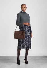 Pulover gri tricotat, fustă din dantelă florală bleu marin și poșetă din suede maro cu închidere aurie. Modelul poartă tocuri negre, arătând ciorapi negri transparenți.