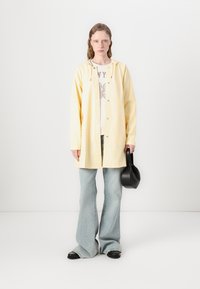 Camisa chaqueta oversized de color amarillo claro con capucha, cierre de botones, llevada sobre una camiseta blanca con gráficos y pantalones vaqueros acampanados de color azul claro. Bolsa negra.