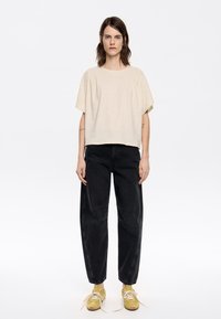 Beige top s krátkym rukávom, voľný strih, skombinovaný s čiernymi džínsami s vysokým pásom a zúženým strihom; model má na sebe žlté tenisky s svetlými šnúrkami.