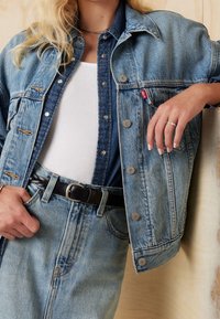 Veste en denim à coupe décontractée, lavage bleu clair, avec des boutons caractéristiques et une étiquette logo rouge. Superposée sur un débardeur côtelé blanc et un denim bleu.