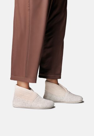 Jambes portant un pantalon marron court et des chaussons en laine gris clair avec des poignets tricotés pliés sur fond blanc.