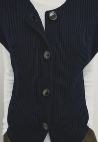 Laivastonsininen neuleliivi, jossa on resorikuosi, syvä V-aukko ja viisi etunappia. Päällä valkoinen pitkähihainen paita, joka korostaa kerrospukeutumista.