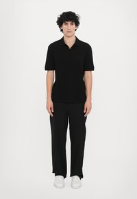 Svart polo skjorte med korte ermer, slim fit, flatt strikket krage. Kombinert med løse svarte bukser og hvite joggesko. Enkelt design, myk tekstur.