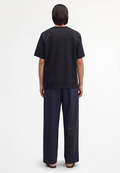 Person mit glatten, dunklen Haaren, der ein schwarzes Kurzarmshirt, dunkle, weit geschnittene Hosen und schwarze Sandalen trägt, steht mit dem Rücken zur Kamera.