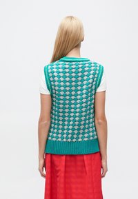 Bezrekawnik dzianinowy z turkusowo-szarym wzorem serc, z prążkowanym kołnierzem i plisą, noszony na białej koszuli i czerwonej spódnicy.