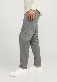 Personne portant un pantalon gris coupe droite, des baskets blanches basses et une chemise à manches longues couleur crème, debout devant un fond uni.