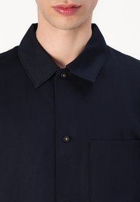 Navyblauwe shirt met een puntkraag, een voorzak en een knoopsluiting. Gemaakt van gestructureerde stof met zwarte knopen.