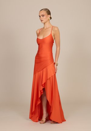Femme en robe orange sans manches avec ourlet haut-bas volanté, boucles d’oreilles dorées, bracelets et talons dorés à fines brides sur fond beige uni.