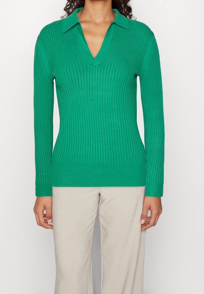 Femme portant un pull ajusté à manches longues en maille côtelée verte avec un col et un décolleté en V, associé à un pantalon beige.