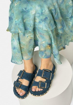 Stopy w granatowych zamszowych sandałach z dużymi klamrami i ozdobnymi podeszwami, zestawione z zwiewną, przezroczystą, niebiesko-zieloną kwiecistą spódnicą.