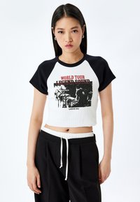 Jonge vrouw met lang zwart haar draagt een zwart-witte cropped raglan T-shirt met de tekst en grafiek "World Tour Legend Sound London 1992".