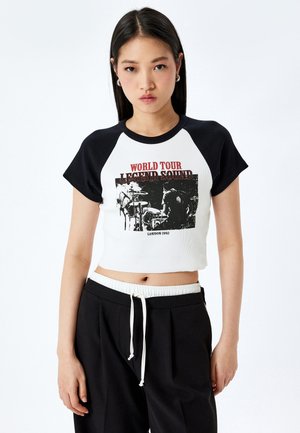 Jonge vrouw met lang zwart haar draagt een zwart-witte cropped raglan T-shirt met de tekst en grafiek "World Tour Legend Sound London 1992".