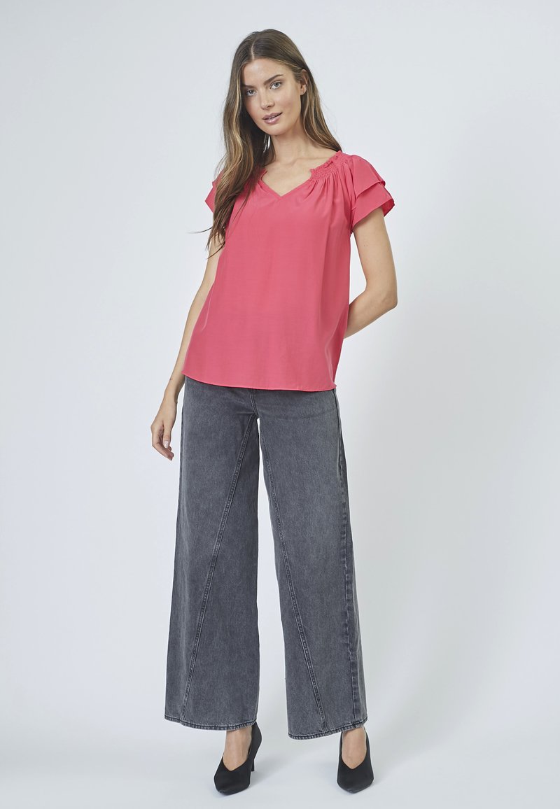 Blusa rosa a maniche corte con scollatura a V, caratterizzata da un design arricciato sulle spalle. Abbinata a jeans grigi scuri a gamba larga e tacchi neri.