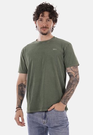 Uomo con capelli ricci che indossa una maglietta verde "TAKE A" e jeans azzurri chiari, che mostra tatuaggi dettagliati su entrambi gli avambracci su sfondo bianco.