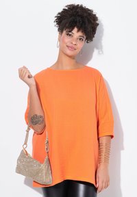 Maglietta oversize arancione e texturizzata con collo a barca, abbinata a leggings neri lucidi. Tiene in mano una borsa metallica dorata. Accenti con bracciale dorato.