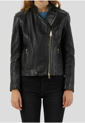 Kobieta nosząca dopasowaną czarną skórzaną kurtkę z asymetrycznym przednim zamkiem błyskawicznym i zamykanymi na suwak bocznymi kieszeniami, w połączeniu z czarnymi spodniami.