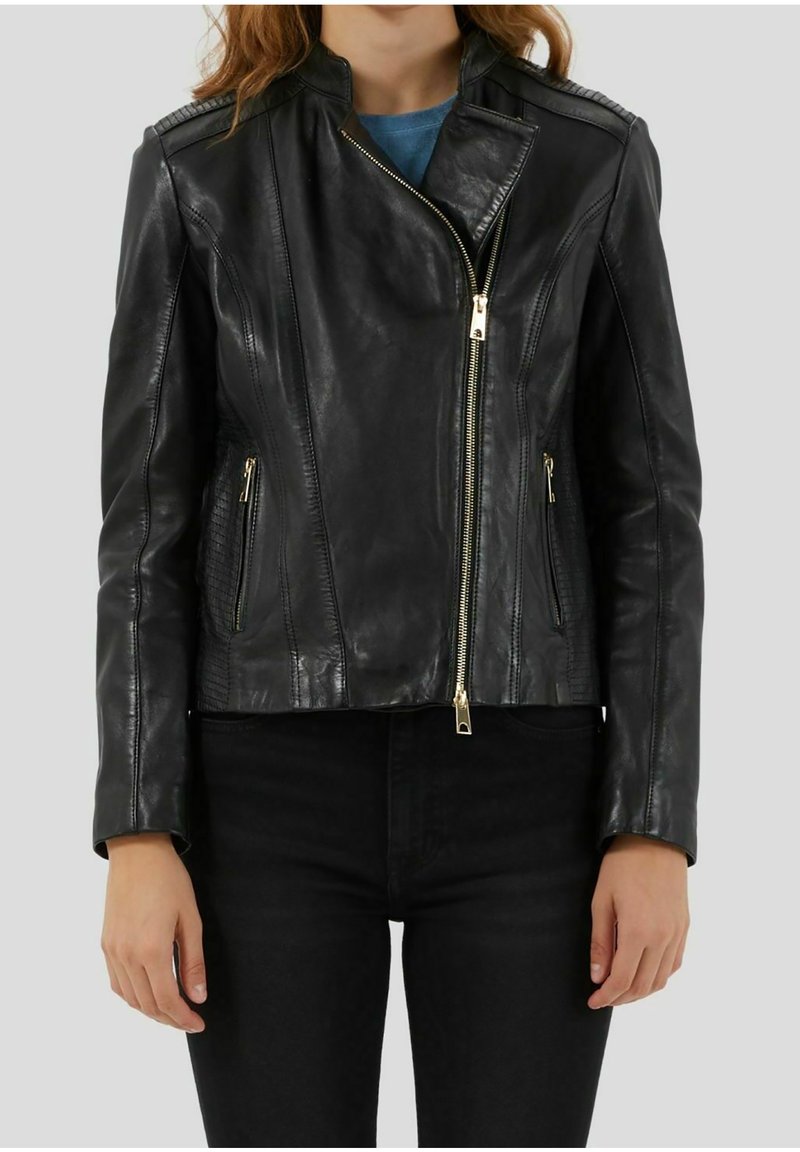 Femme portant une veste en cuir noire cintrée avec une fermeture éclair asymétrique à l'avant et des poches latérales zippées, associée à un pantalon noir.