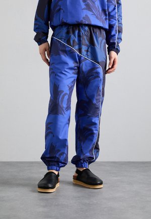 Blue track pants featuring a palm leaf pattern in black, elastic cuffs, and a contrasting black side panel with white piping. Black clogs with wooden soles.