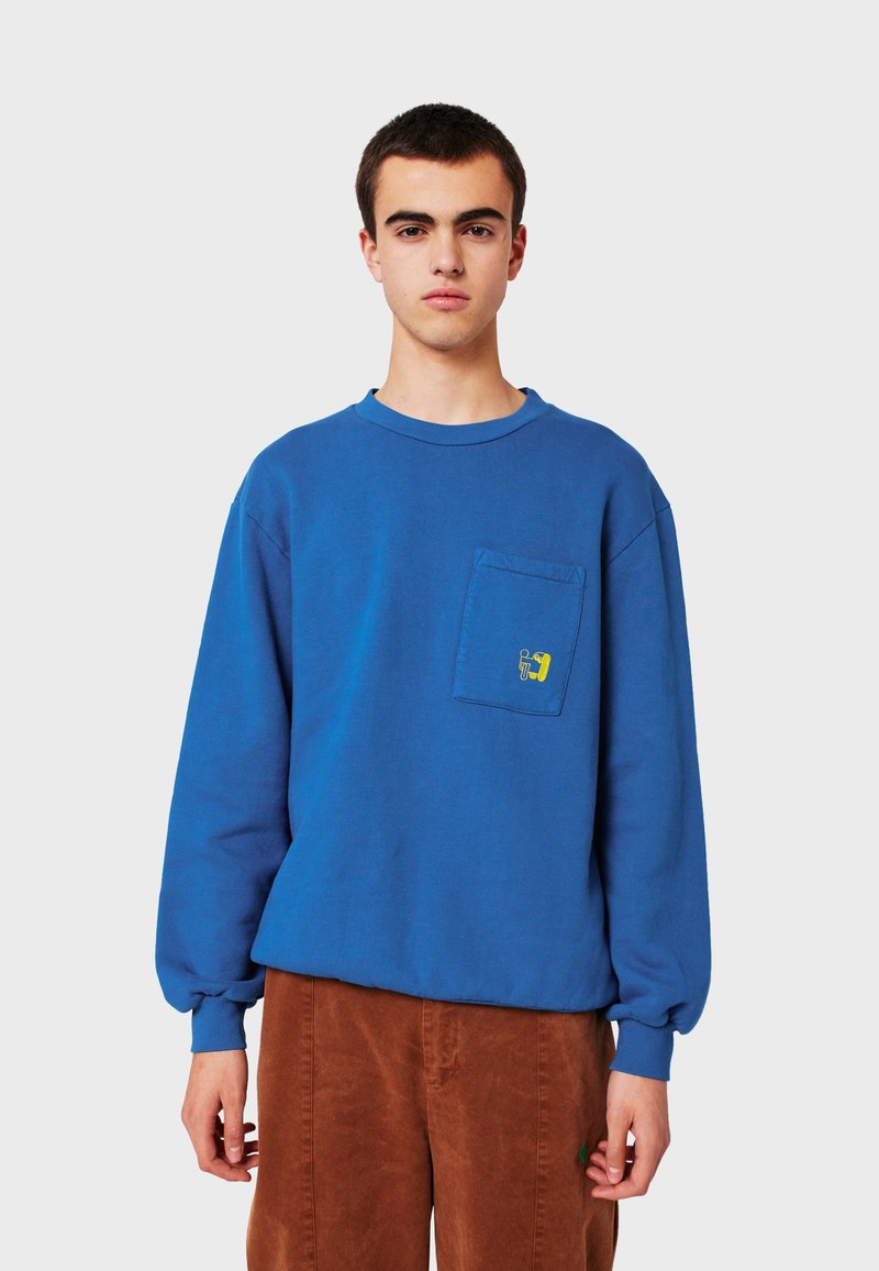 Joven con una camisa de manga larga azul con un gráfico amarillo en el bolsillo del pecho y pantalones de pana marrones, de pie contra un fondo gris.