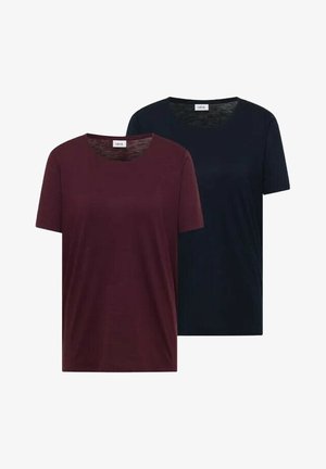 Junge Frau mit langen braunen Haaren, die ein marineblaues kurzärmeliges T-Shirt und hellblaue Jeans trägt, mit den Händen in den Taschen.