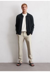 Black button-up cardigan over a white t-shirt, paired with light beige trousers and black sandals. Minimalist design, smooth fabric textures.